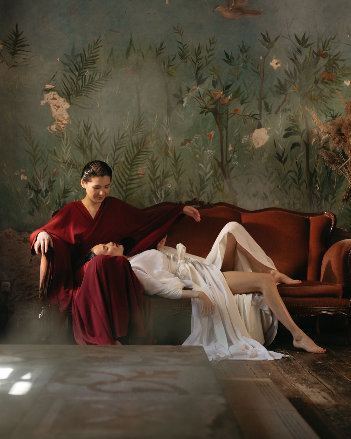 Two women resting on a velvet sofa in front of a painted mural.