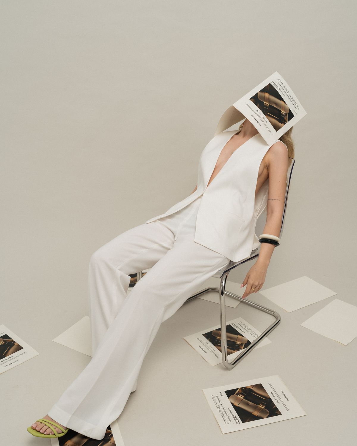 Model in a white suit seated on a metal chair against a pale wall.